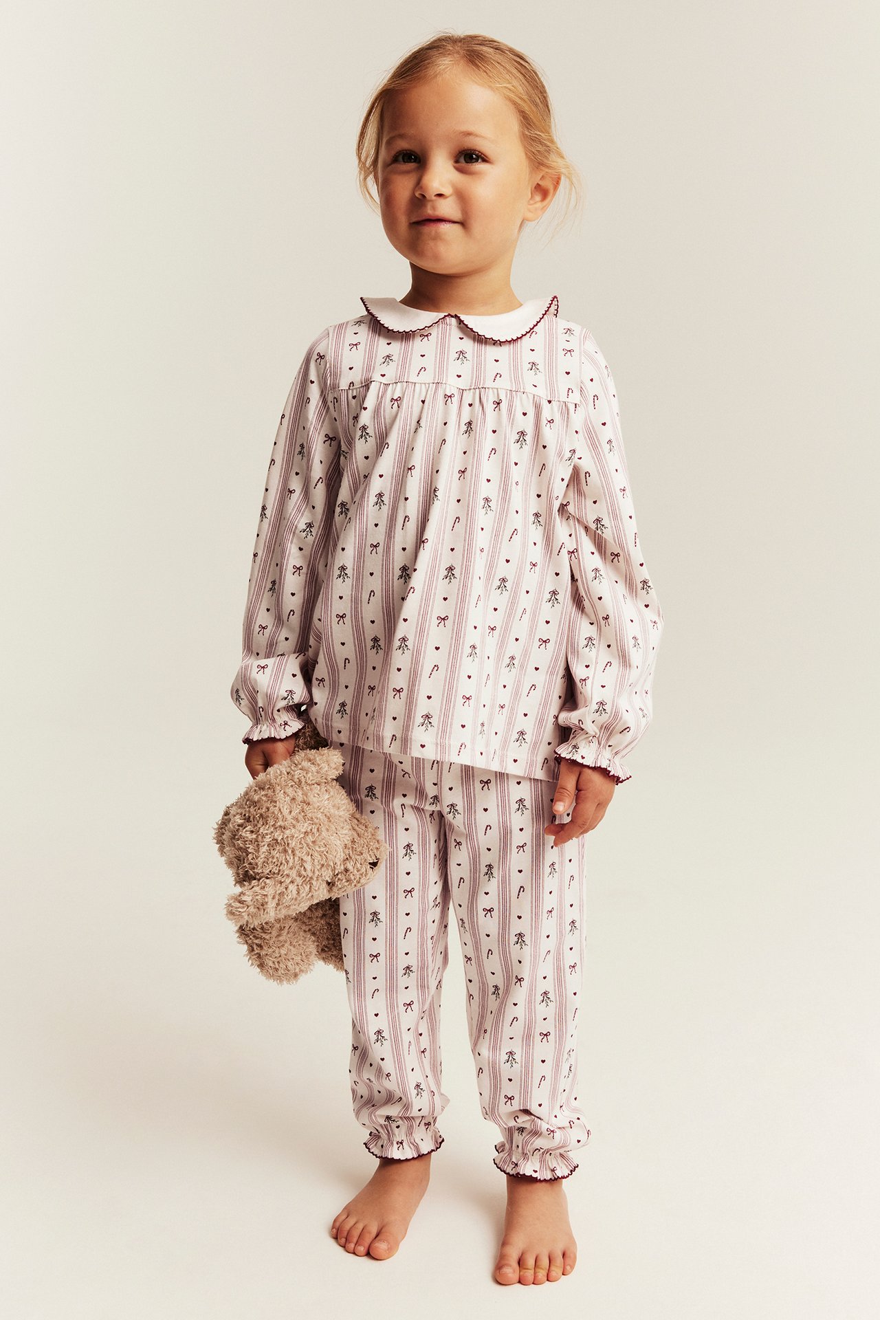 Framsida: Barnpyjamas vit-vinröd randig med mönster, barn håller nallebjörn.