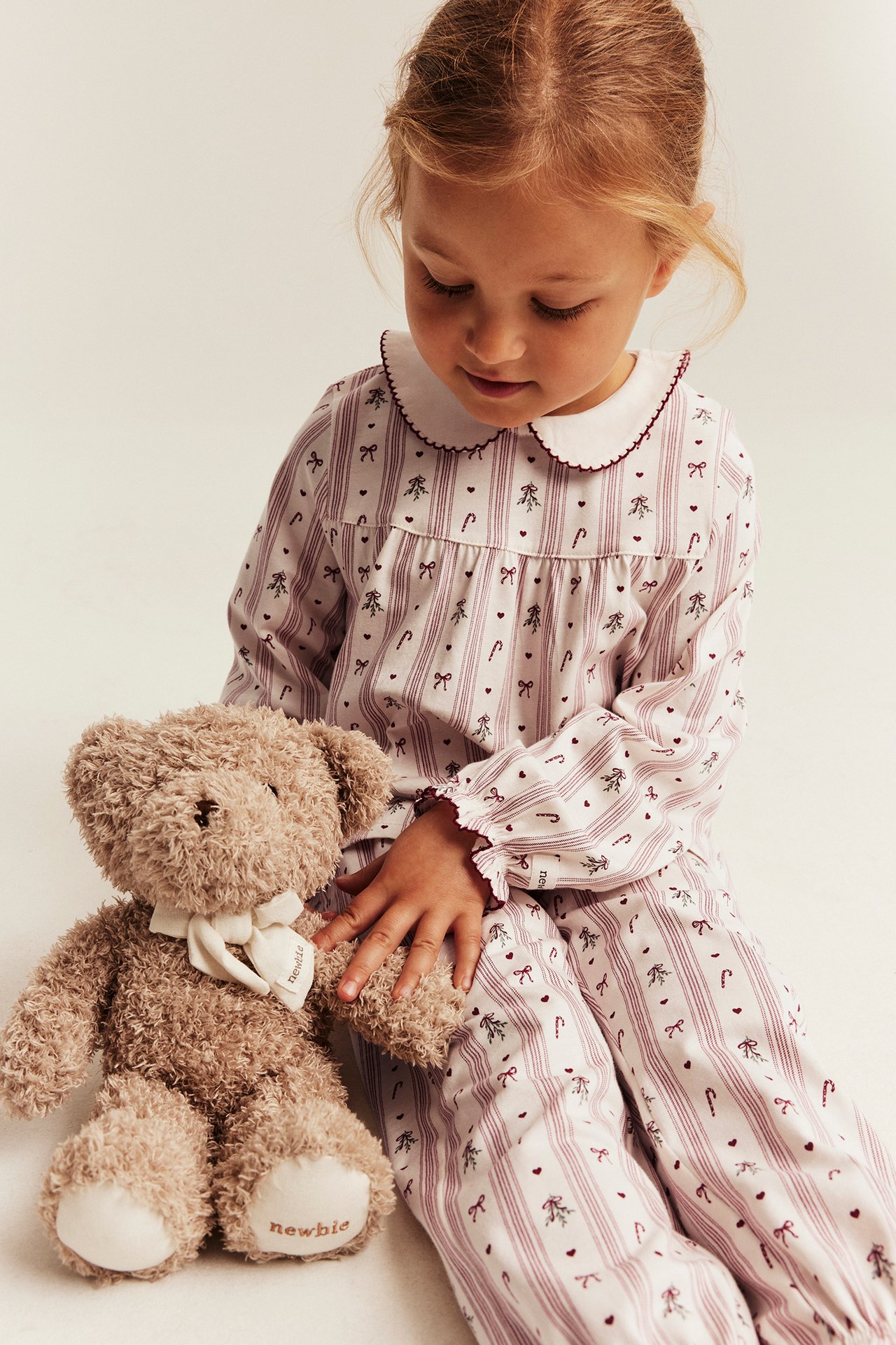 Framifrån, barn i vit pyjamas med röda ränder, rosetter, polkagrisar, håller i brun nallebjörn.