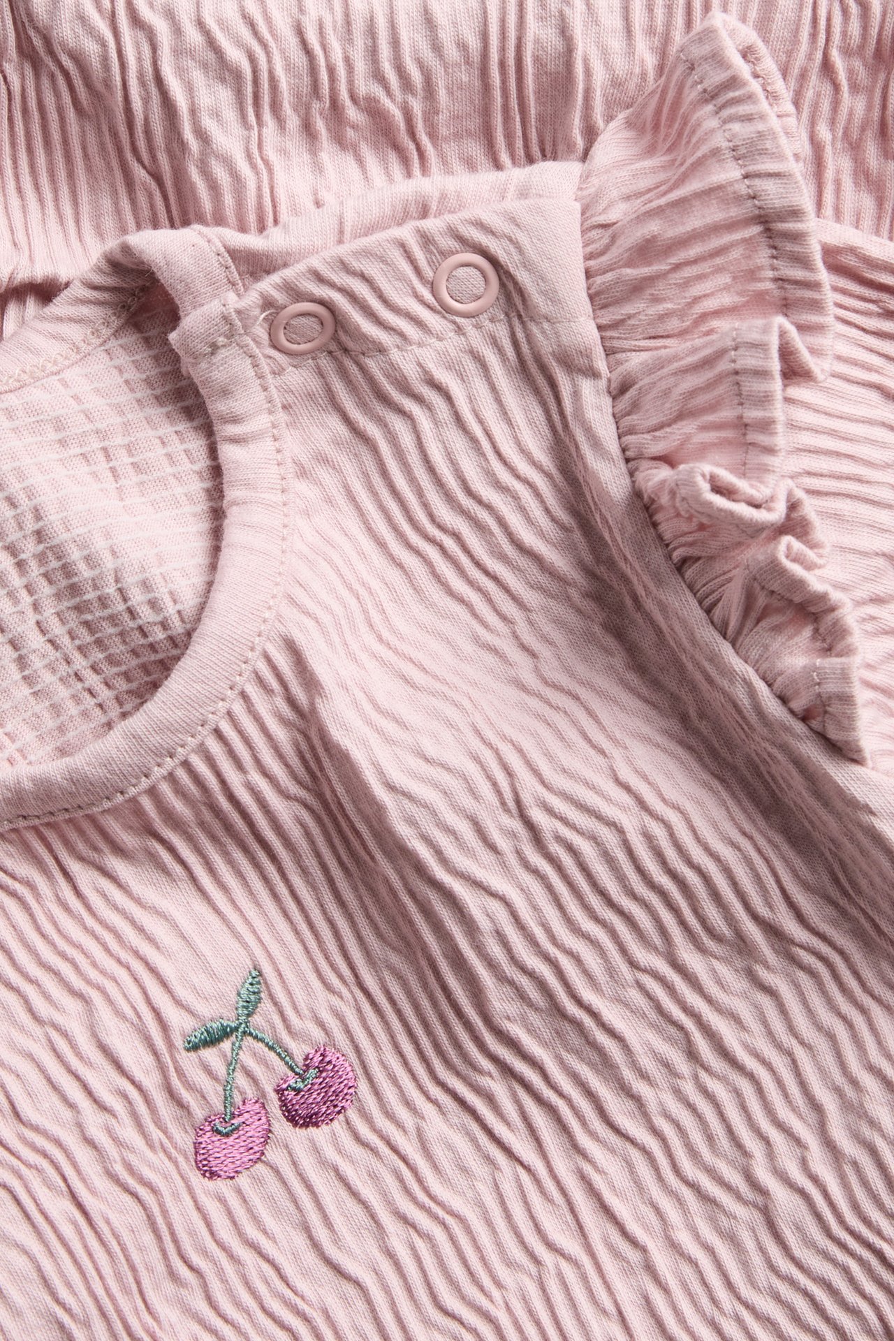Nærbilde av rosa krøllete babyplagg med skulderknapper, volang og kirsebærbroderi.