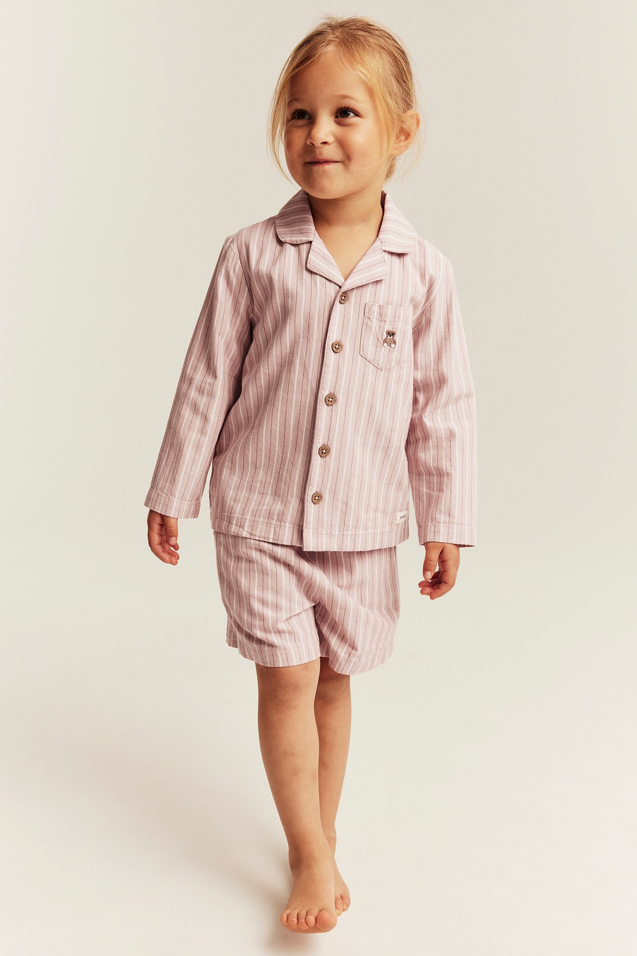 Frontvy: Barn i rosa randigt pyjamasset, långärmad tröja med björnbroderi och shorts.