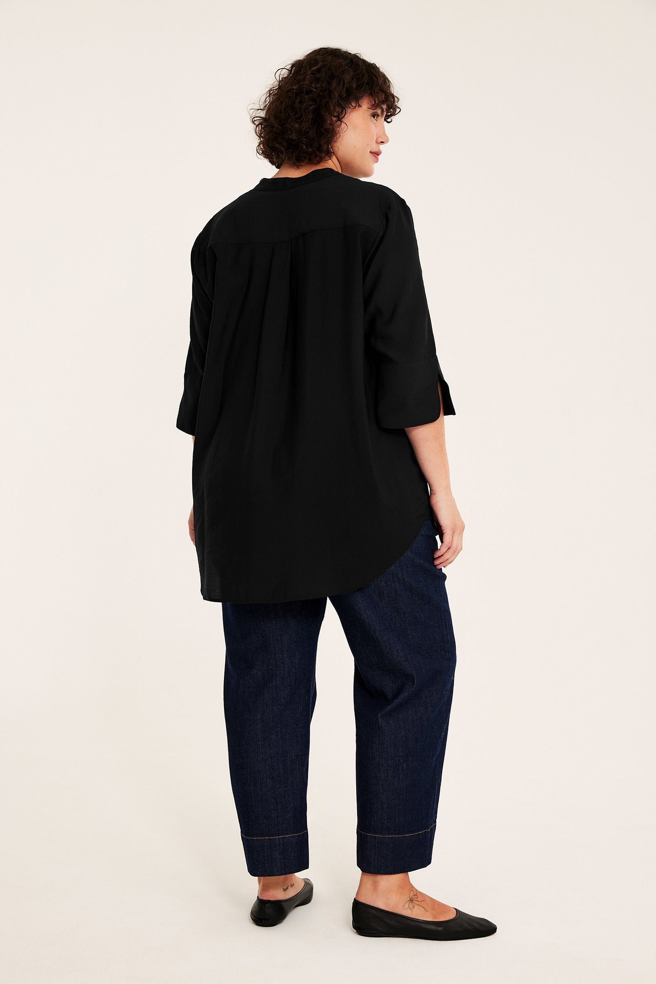 Bakfra av kvinnelig modell i svart tunikaskjorte, mørkeblå jeans og svarte ballerinasko.