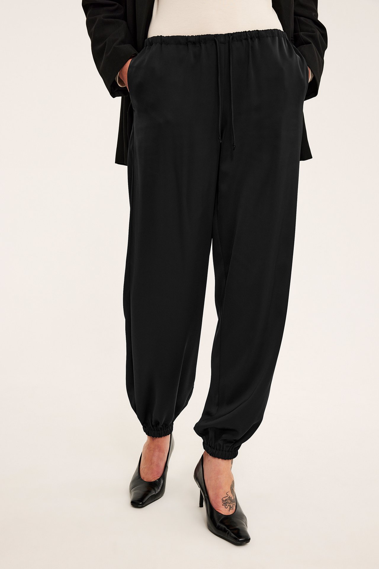 Framifrånbild på kvinna med svarta satinjoggers, krämfärgad topp, svart kavaj och pumps.