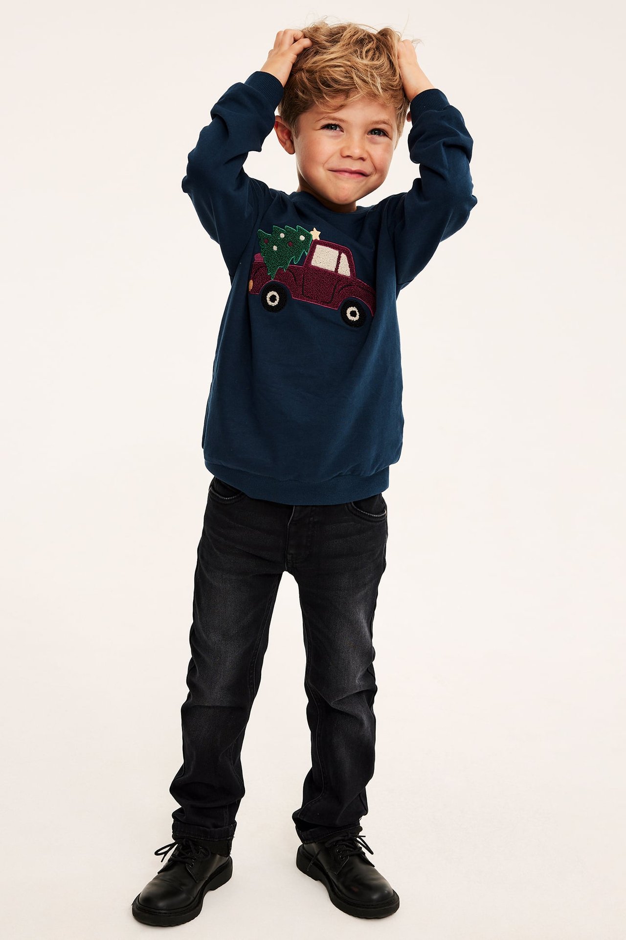 Frontvy av ett barn i mörkblå sweatshirt med röd lastbil och julgran, svarta jeans och svarta skor.