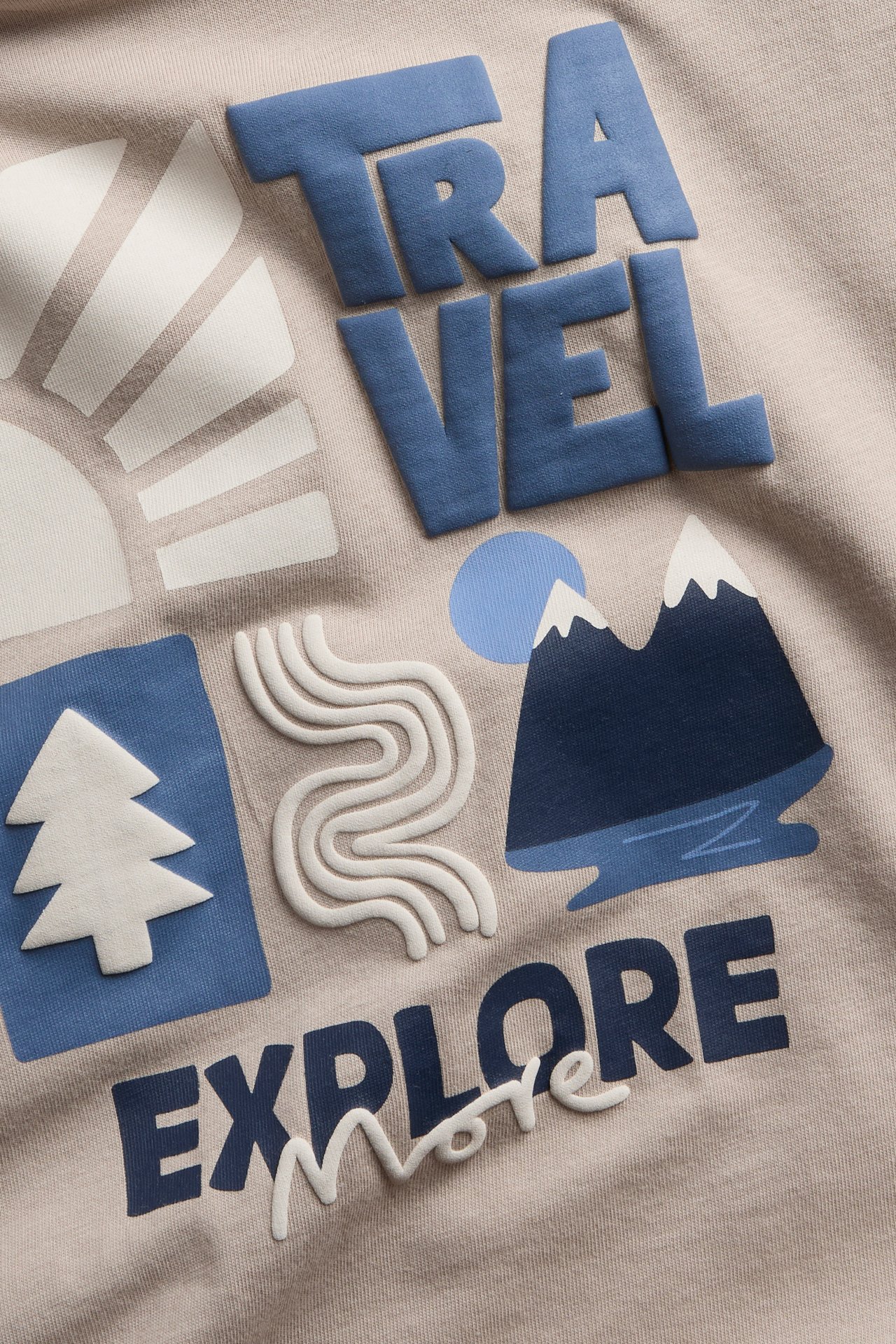 Zbliżenie jasnobeżowej bluzki z niebiesko-białym, wieloteksturowym nadrukiem „TRAVEL EXPLORE more”.