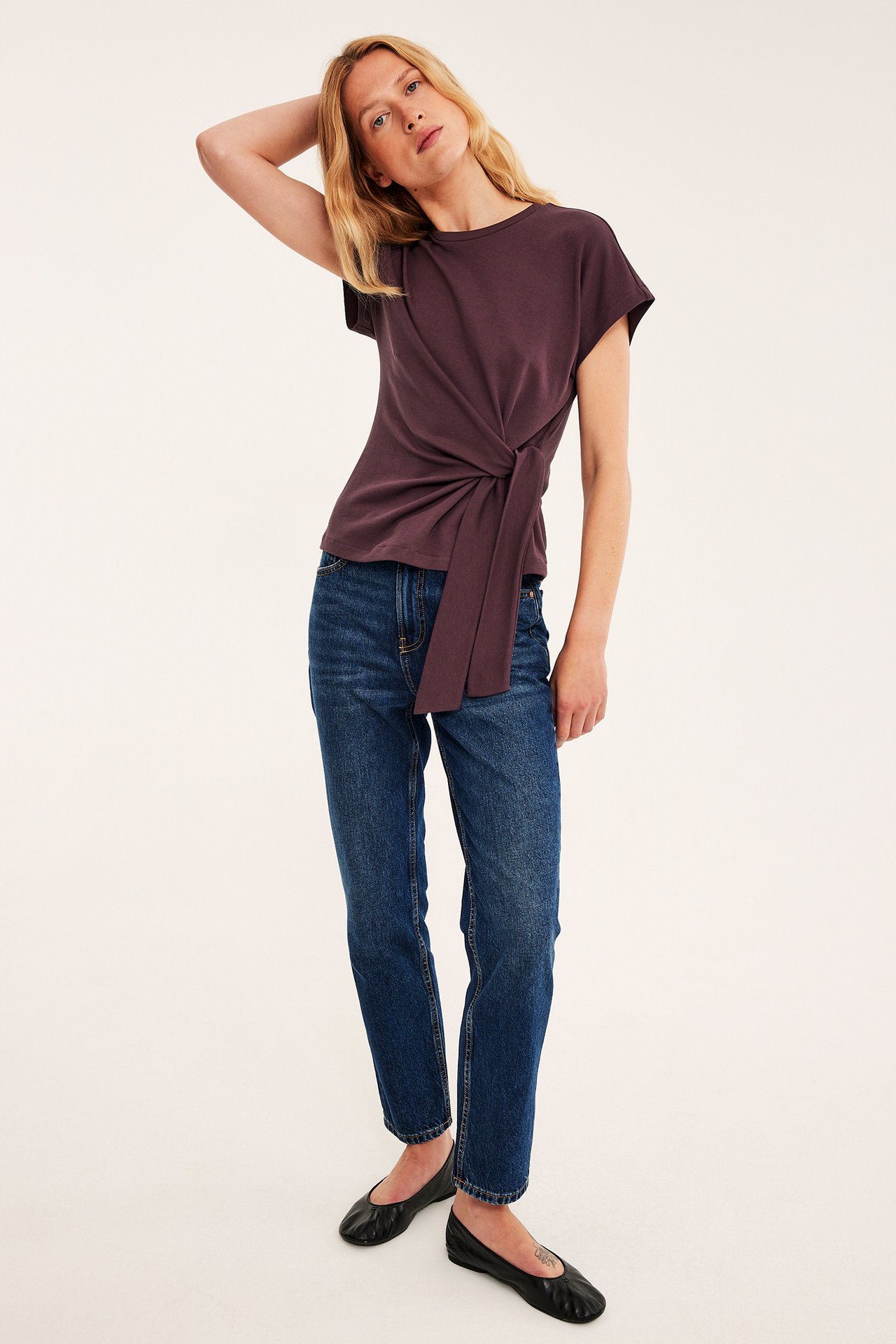 Framifrån en kvinna i mörk lila knuten t-shirt, blå jeans och svarta ballerinaskor.