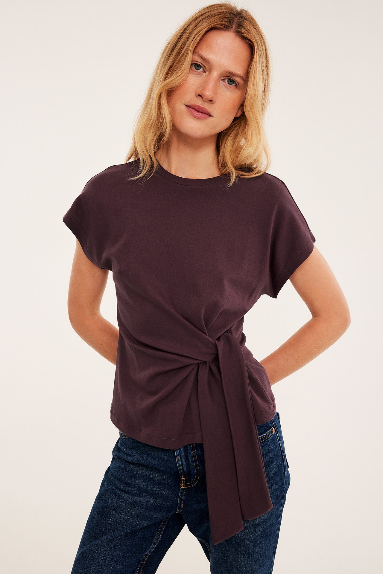 Framifrån, kvinnlig modell i mörklila kortärmad t-shirt med sidoknut och mörkblå jeans.