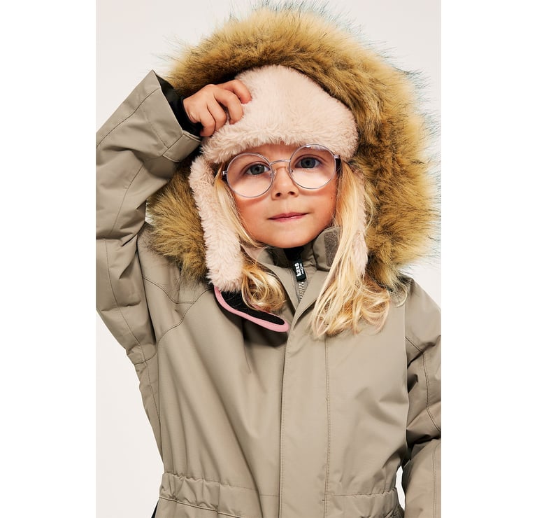 Flicka i beige vinteroverall med luva i fuskpäls och fleecefodrad mössa.