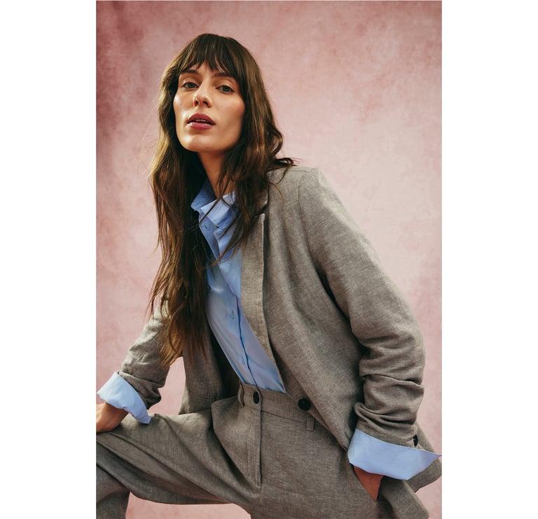 Kobieta w lnianym garniturze siedząca na krześle na różowym tle.