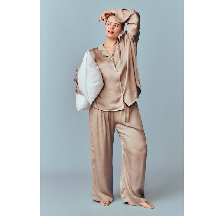 Kobieta stojąca w kremowej piżamie, trzymająca poduszkę.