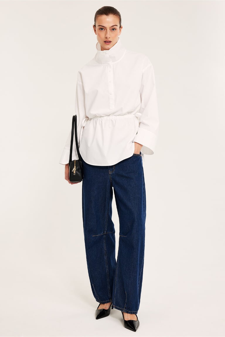 Kvinne i hvit skjorte med høy krage, tynne jeans og svart veske.
