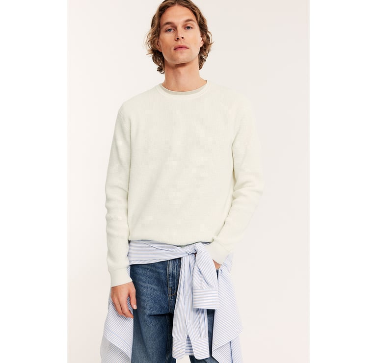 Mężczyzna w białym swetrze, dżinsach i niebieskiej koszuli zawiązanej w talii.