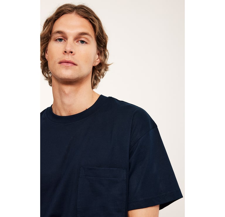 Mężczyzna w białym swetrze, dżinsach i niebieskiej koszuli zawiązanej w talii.