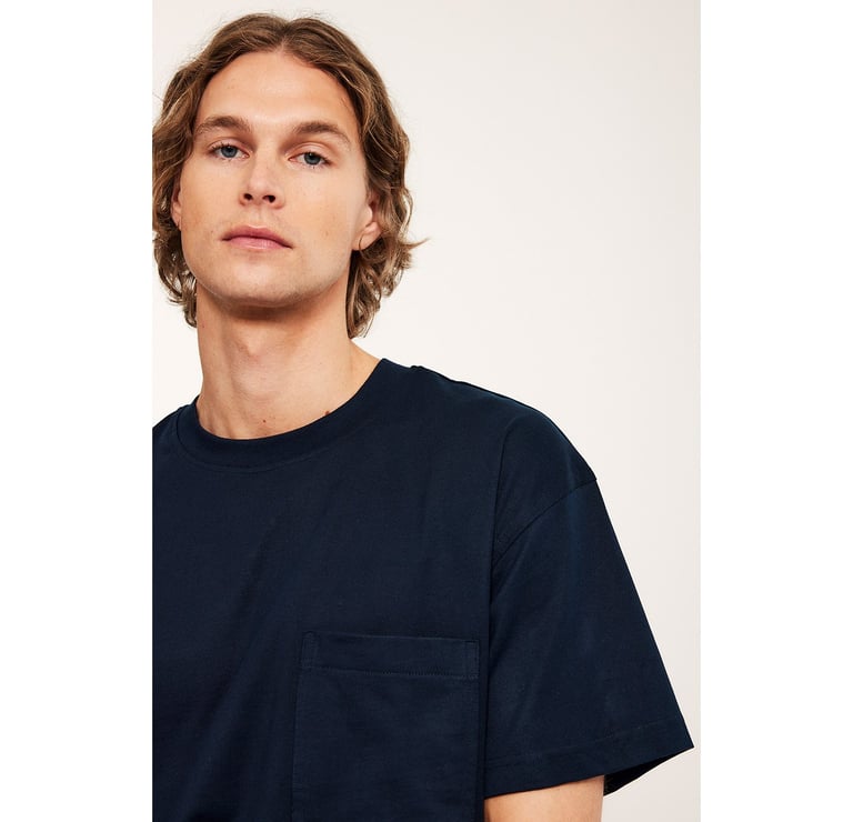 Mężczyzna w białym swetrze, dżinsach i niebieskiej koszuli zawiązanej w talii.