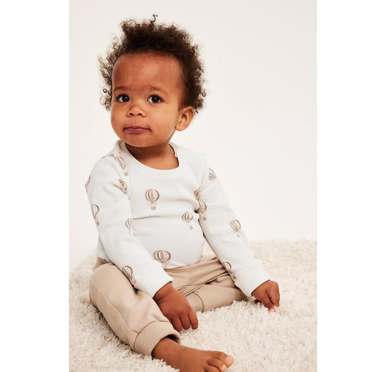 Niemowlę w białym body z balonikami i beżowych spodniach dresowych.