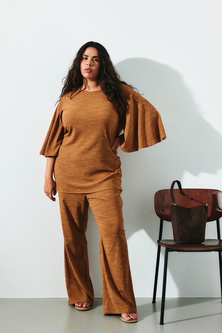 Plus size kläder till kvinnor. Kvinna i matchande set i guld på vida byxor och topp med vida ärmar.