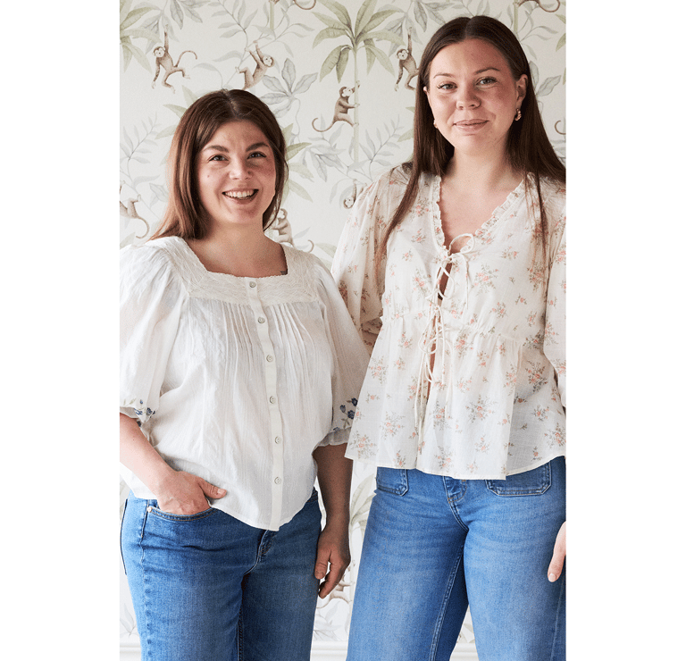 Kaksi ruskeatukkaista naista seisoo tapetoidun seinän edessä puseroissa ja farkuissa.