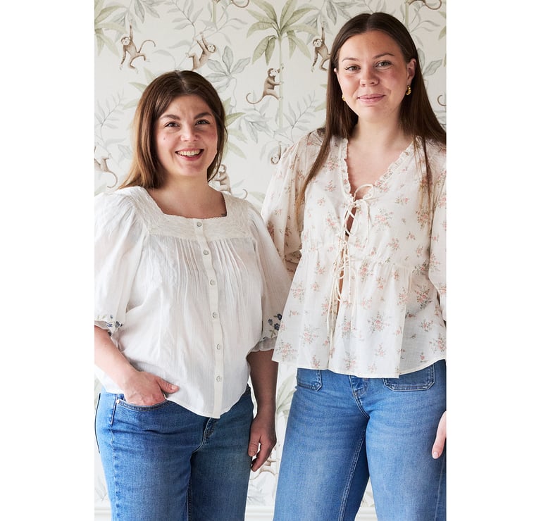 Kaksi ruskeatukkaista naista seisoo tapetoidun seinän edessä puseroissa ja farkuissa.